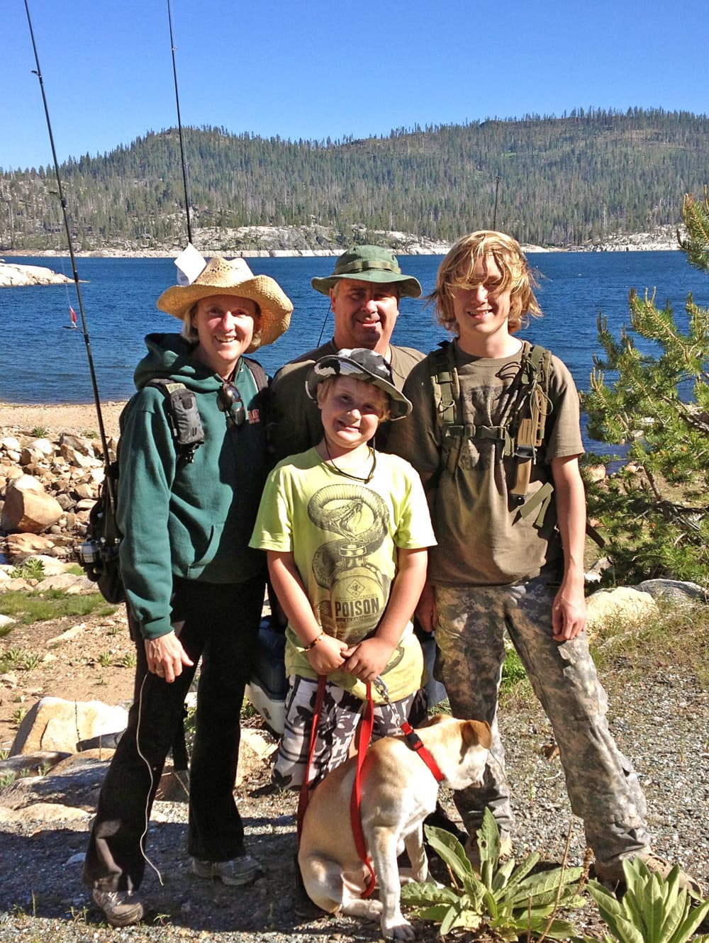 A family of four stands by a lake, enjoying a sunny day with fishing rods in the background.
