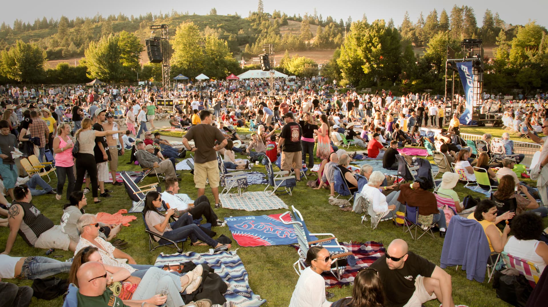 A large crowd gathers on a grassy field for an outdoor event, surrounded by trees and tents.