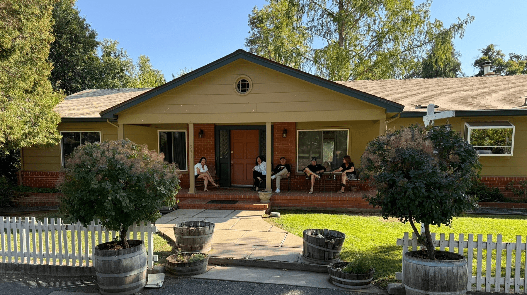 A group of five people sits on the porch of a yellow house surrounded by greenery.
