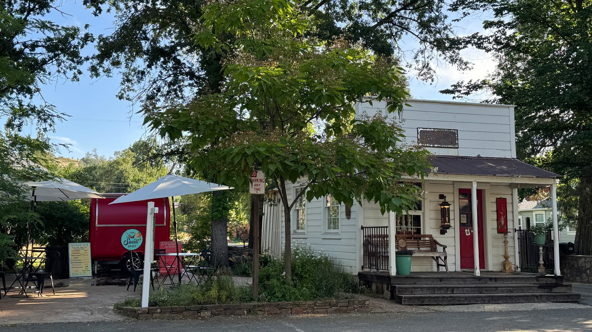 A mini white wooden two-story house with a small porch and trees around it and a small food trailer beside it with a seating area.