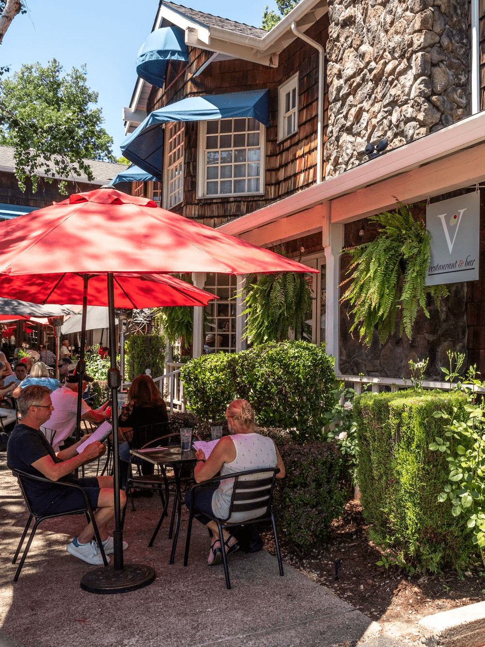 Outdoor dining scene at a restaurant featuring red umbrellas and patrons enjoying their meals.