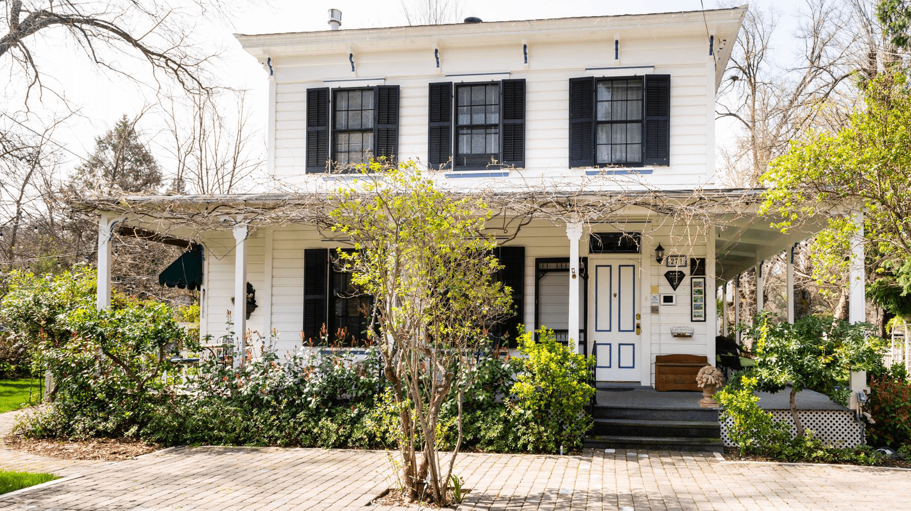 A charming white two-story house with black shutters, surrounded by greenery and a brick walkway.