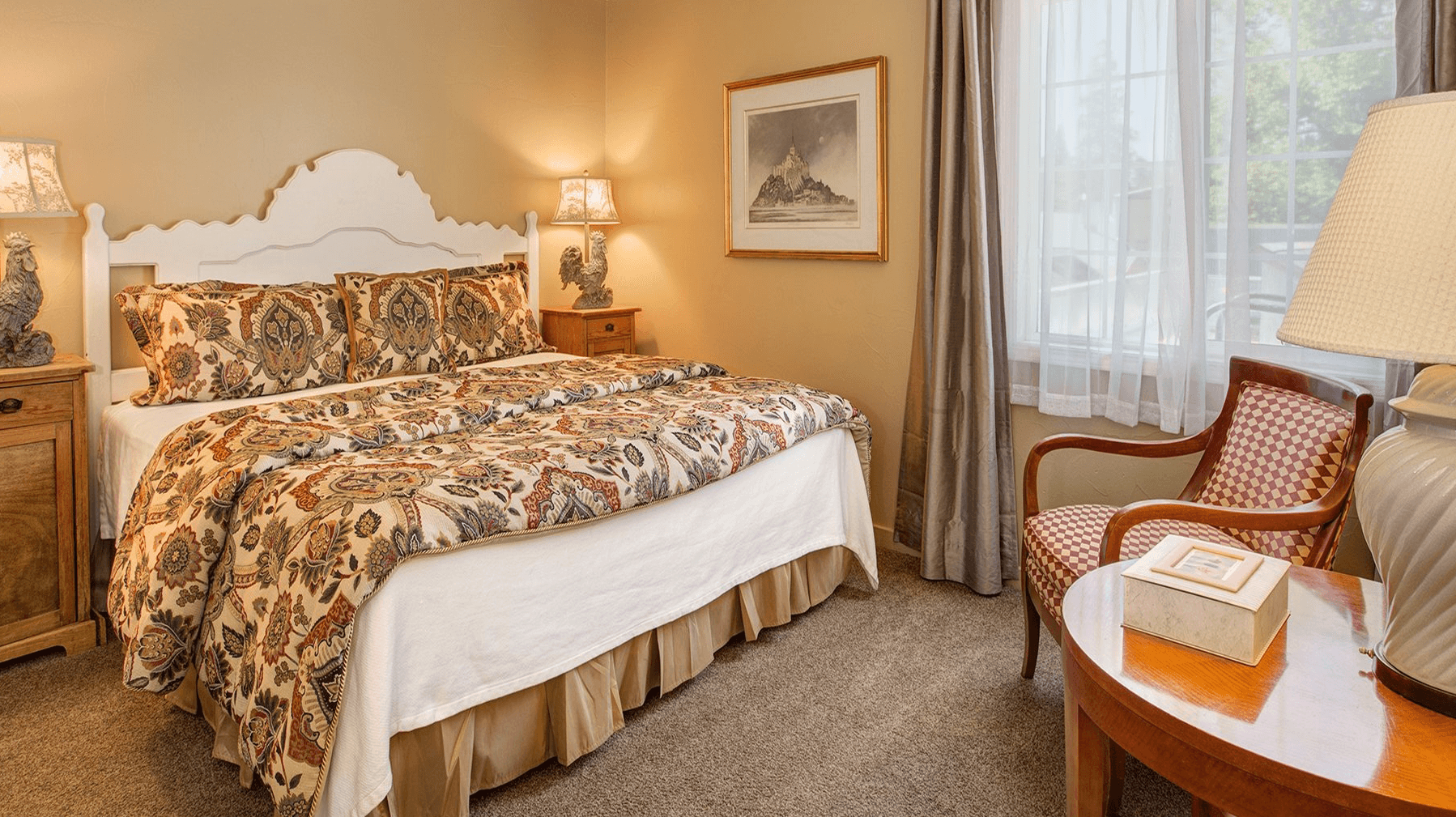 Cozy bedroom featuring a large bed with patterned bedding, a wooden nightstand, and a chair beside a window.