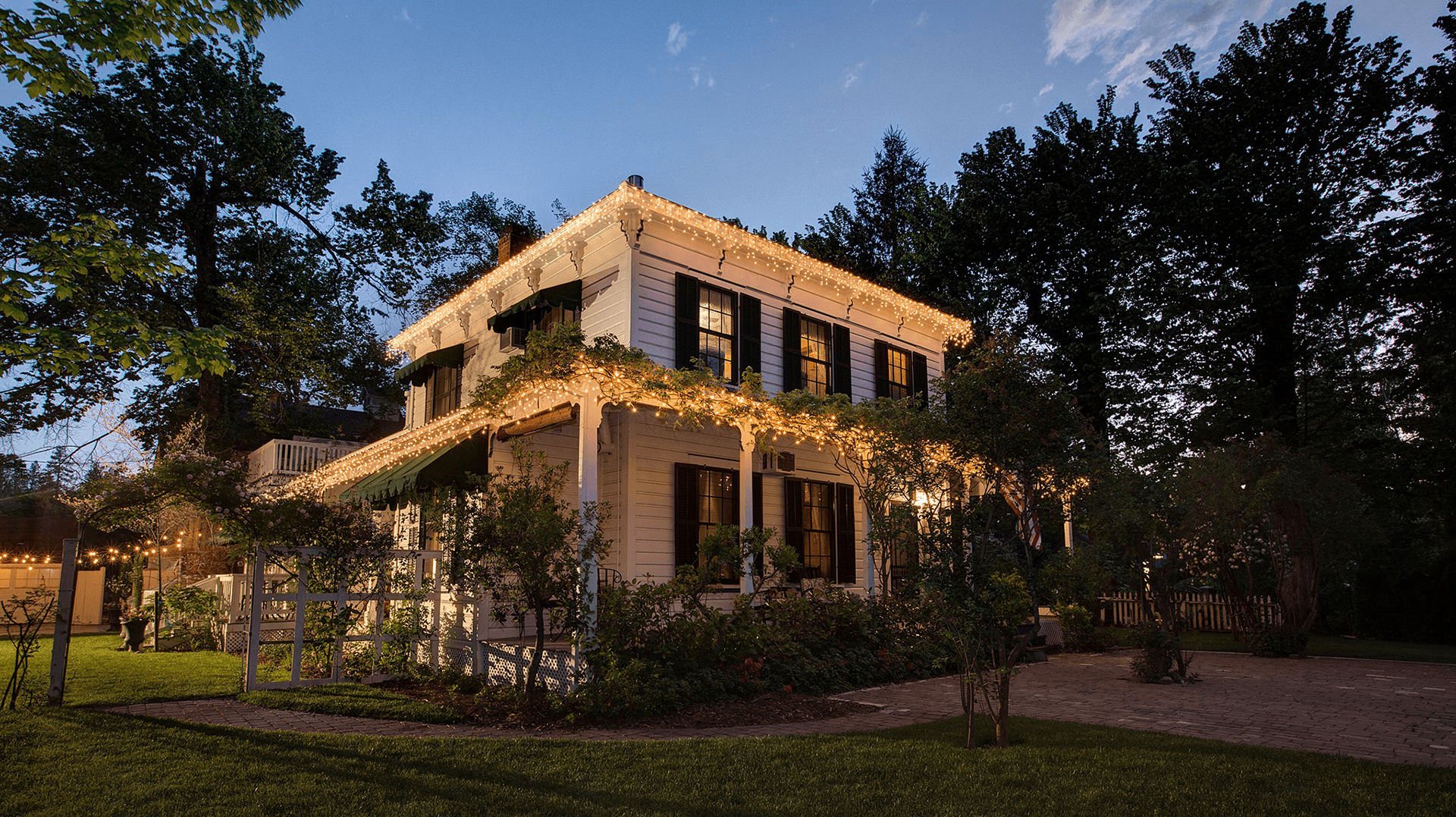 A charming white house adorned with warm lights, surrounded by lush greenery at dusk.